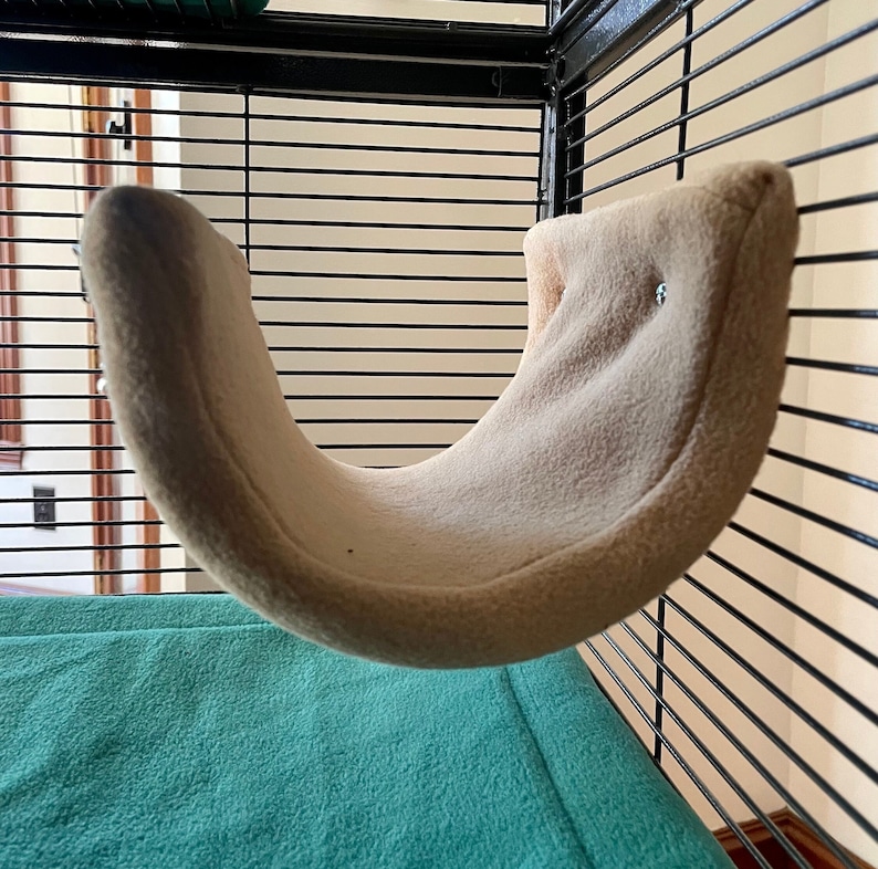 May include: A beige hammock-shaped bed for small pets, hanging inside a black wire cage. The bed is made of soft, plush fabric and has a curved shape.