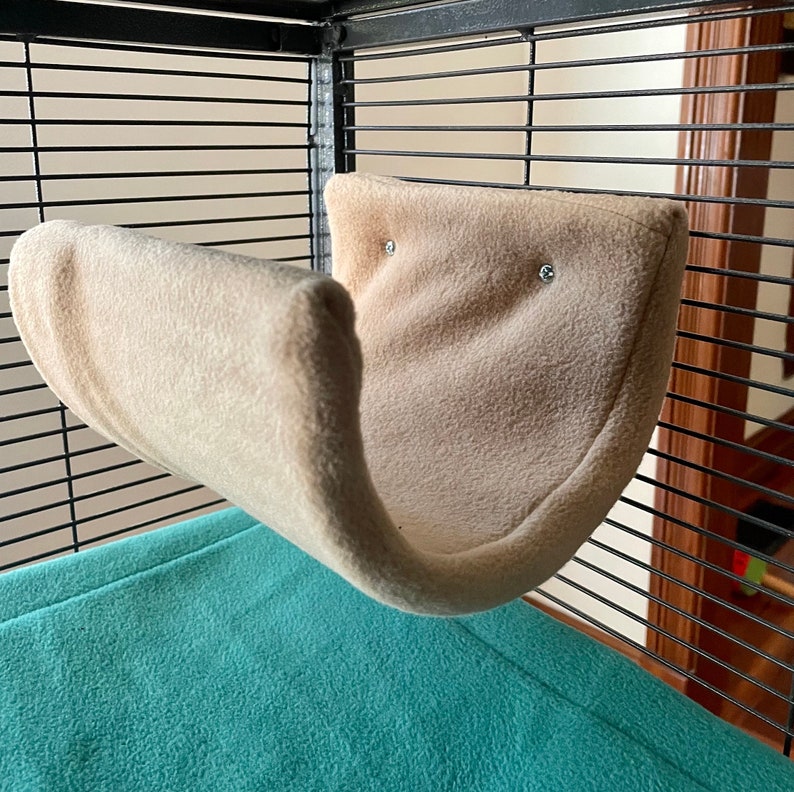 May include: A beige fleece hammock for small animals, hanging inside a cage with a teal blue fleece liner on the bottom.