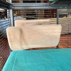May include: Two beige fleece hammock-style hanging beds for small animals inside a cage with a teal blue fleece bottom.