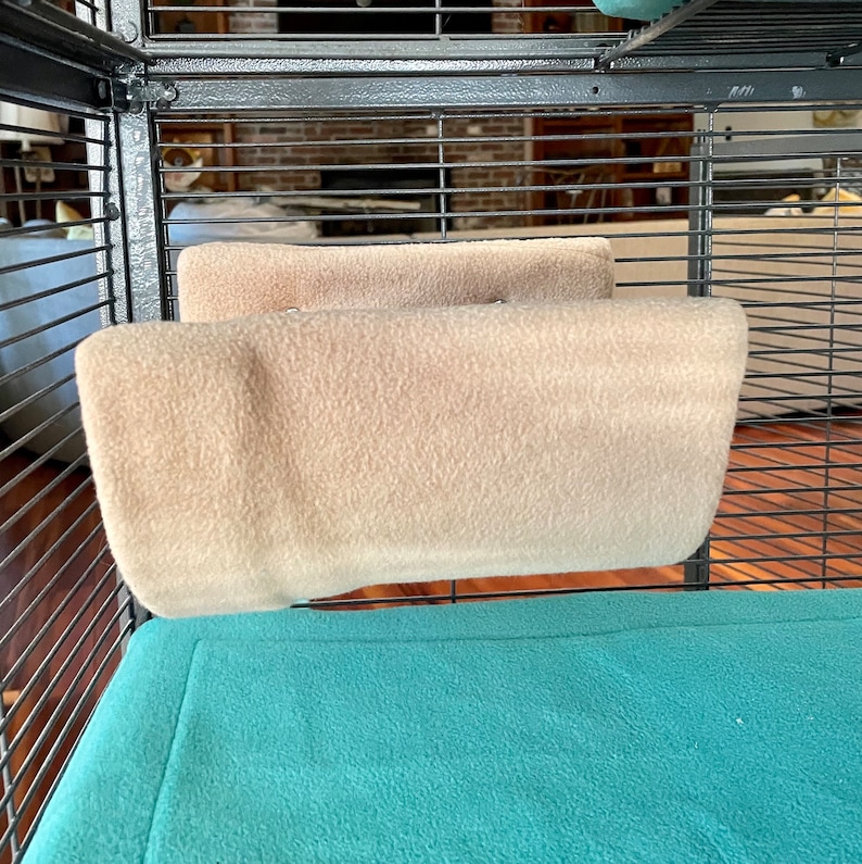 May include: Two beige fleece hammock-style hanging beds for small animals inside a cage with a teal blue fleece bottom.