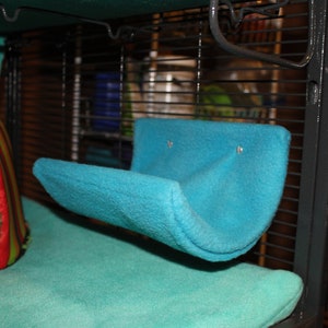 May include: A turquoise blue fleece hammock for small animals, hanging inside a black wire cage. The hammock is attached to the cage with metal hooks.