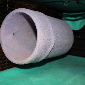 May include: A lavender-colored, cylindrical pet tunnel made of soft, textured fabric. The tunnel is suspended inside a wire cage, with a green mat visible below. A small metal fastener secures the tunnel.