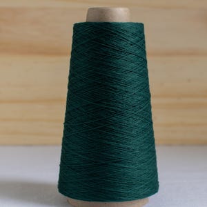May include: A spool of dark green yarn, cone-shaped, with a wooden top and base. The yarn is tightly wound, creating a textured surface. The background is a light wooden panel.