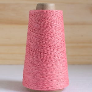 May include: A spool of coral-colored thread, tightly wound on a cardboard core. The thread is a solid color, and the spool is set against a light wooden background. The thread is likely used for sewing, embroidery, or other textile crafts.
