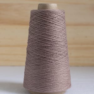 May include: A spool of taupe-colored yarn, cone-shaped, with a cardboard core. The yarn is tightly wound, creating a textured surface. The background is a light wooden panel and a white surface.