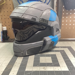 May include: A gray and blue futuristic helmet with a dark visor. The helmet has a segmented design with blue accents and a glossy finish. The helmet is sitting on a gray and white checkered surface.