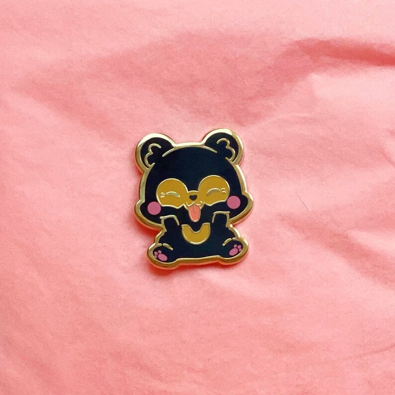 Puede incluir: Un pin de esmalte negro con un oso de dibujos animados que lleva un contorno dorado. El oso tiene una cara amarilla, mejillas rosadas y una lengua grande y rosa que sobresale. El oso est&aacute; sentado con las patas delante.