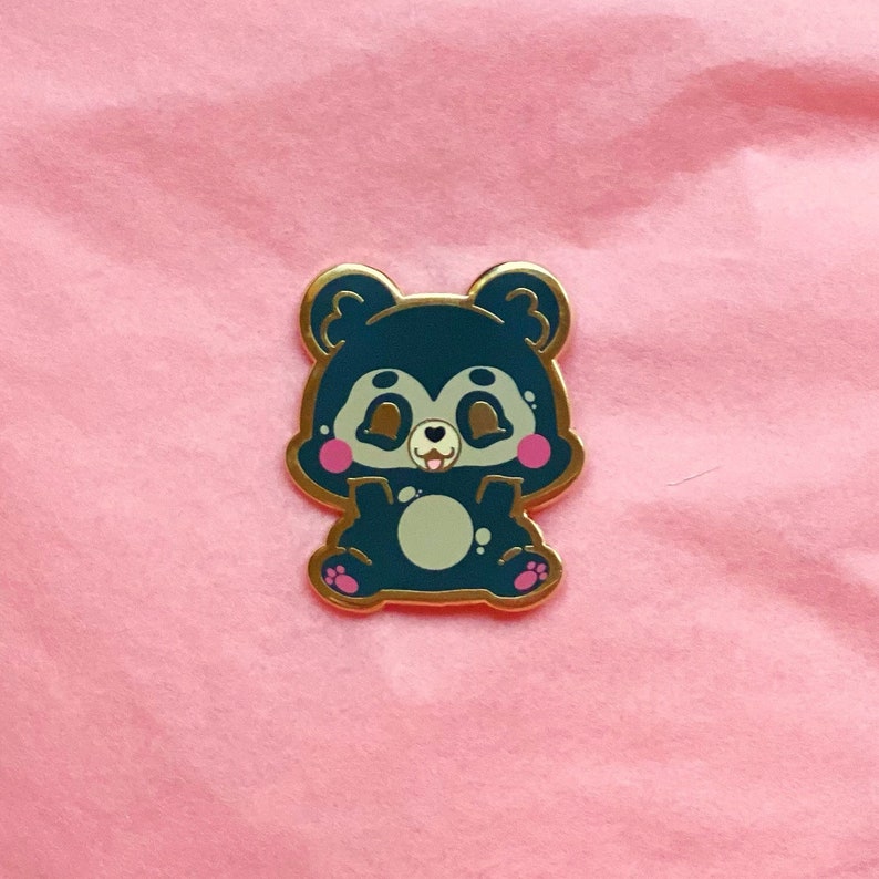 Puede incluir: Un pin de esmalte azul y dorado con un oso de dibujos animados con mejillas rosadas y un vientre verde claro. El oso est&aacute; sentado con las patas delante.