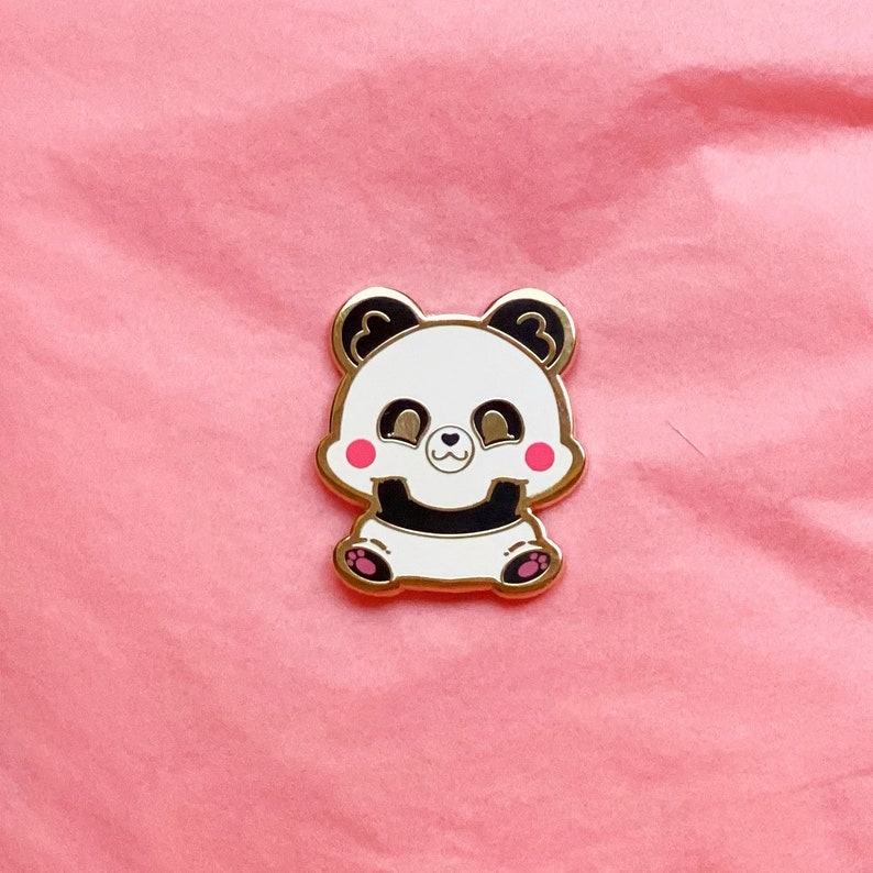 Puede incluir: Pin de esmalte dorado con un panda de dibujos animados con ojos negros, nariz negra y mejillas rosadas. El panda est&aacute; sentado con las patas delante.