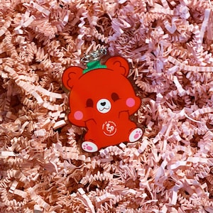 Pode incluir: Chaveiro de urso de desenho animado vermelho com um chap&eacute;u verde e um cora&ccedil;&atilde;o branco no peito. O urso est&aacute; segurando suas patas na frente dele. O chaveiro tem um pequeno logotipo de galo branco com o texto "Home of Zhima" nele.