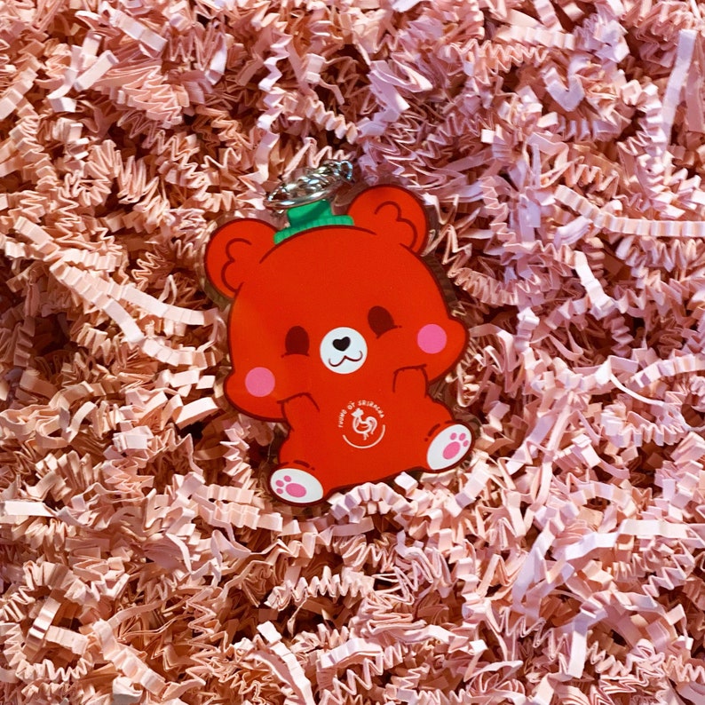 Pode incluir: Chaveiro de urso de desenho animado vermelho com um chap&eacute;u verde e um cora&ccedil;&atilde;o branco no peito. O urso est&aacute; segurando suas patas na frente dele. O chaveiro tem um pequeno logotipo de galo branco com o texto "Home of Zhima" nele.