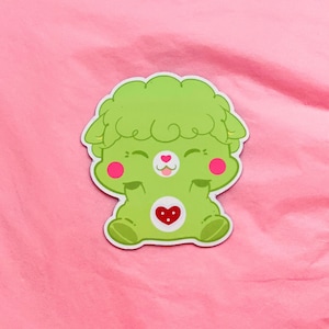 K&ouml;nnte beinhalten: Gr&uuml;ner Care Bear mit einem Herzen auf der Brust und einem flauschigen gr&uuml;nen Kopf. Der Care Bear hat rosa Wangen und einen fr&ouml;hlichen Ausdruck.