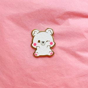 Puede incluir: Un pin de esmalte blanco con acabado brillante que representa un oso de dibujos animados con mejillas rosadas y un ojo gui&ntilde;ando. El oso est&aacute; sentado con las patas delante de &eacute;l.