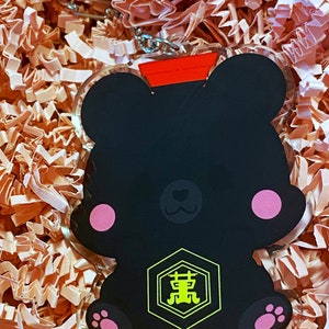 Pode incluir: Chaveiro em forma de urso preto com uma tampa vermelha e um amuleto hexagonal amarelo com caracteres japoneses. O amuleto tem um contorno preto e os caracteres est&atilde;o em amarelo.