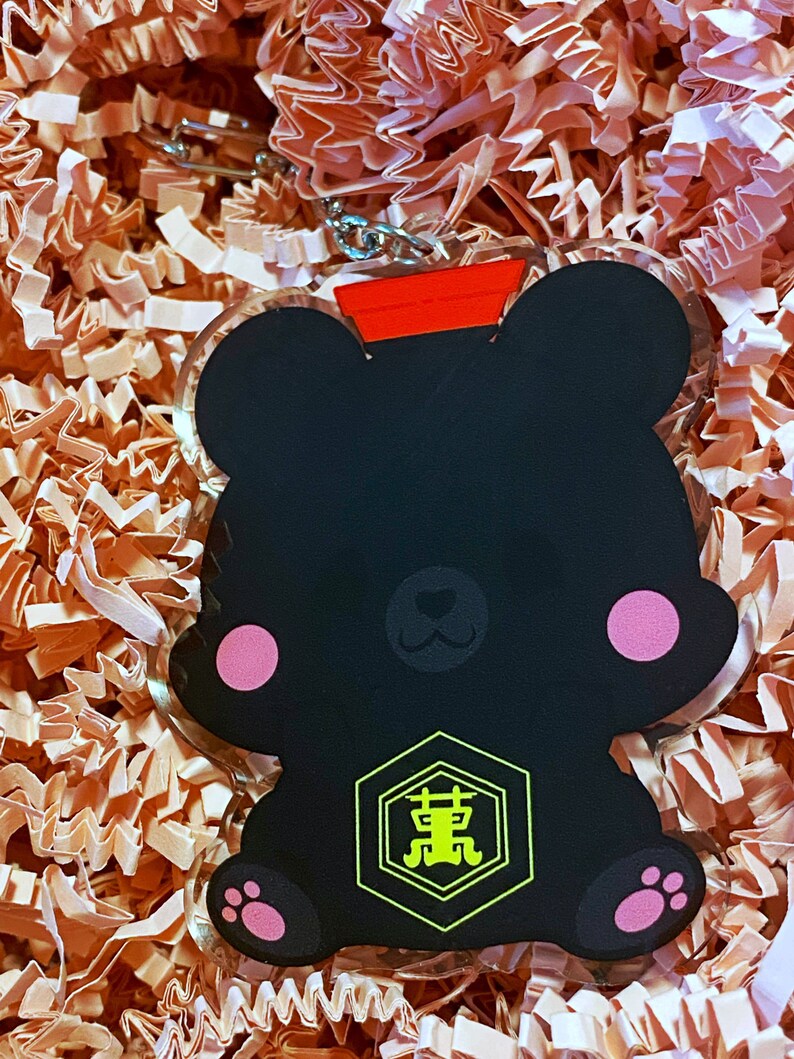Pode incluir: Chaveiro em forma de urso preto com uma tampa vermelha e um amuleto hexagonal amarelo com caracteres japoneses. O amuleto tem um contorno preto e os caracteres est&atilde;o em amarelo.