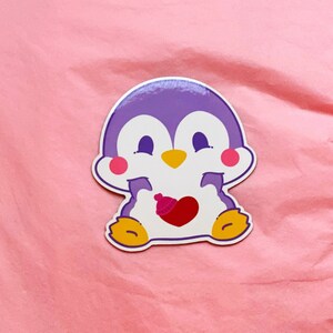K&ouml;nnte beinhalten: Ein lila Pinguin-Aufkleber mit einem rosa Herzen und einem roten Hut. Der Pinguin l&auml;chelt und h&auml;lt das Herz in seinen Fl&uuml;geln.