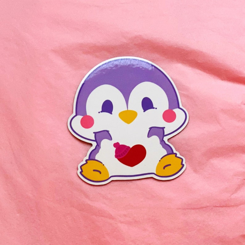 K&ouml;nnte beinhalten: Ein lila Pinguin-Aufkleber mit einem rosa Herzen und einem roten Hut. Der Pinguin l&auml;chelt und h&auml;lt das Herz in seinen Fl&uuml;geln.