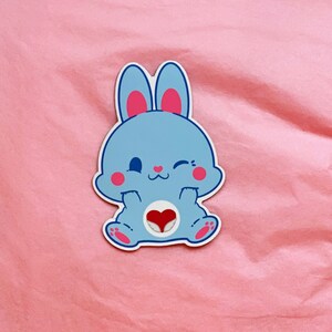 K&ouml;nnte beinhalten: Ein blauer Hasenaufkleber mit einem rosa Herzen auf der Brust. Der Hase hat ein zwinkerndes Auge und ein geschlossenes Auge. Der Aufkleber befindet sich auf einem rosa Hintergrund.