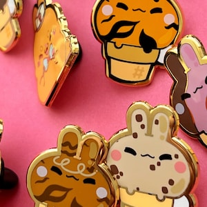 May include: A set of gold-tone enamel pins featuring cartoon ice cream cones with bunny ears. The ice cream cones are decorated with different patterns and colors, including brown, orange, and white. The pins are perfect for adding a touch of whimsy to your clothing or bag.