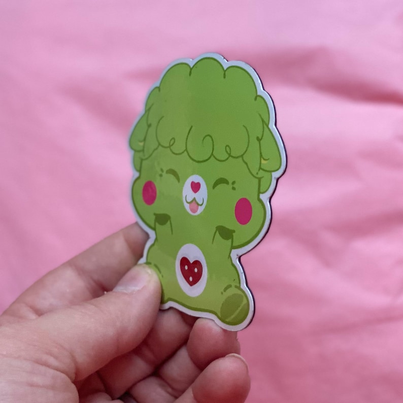 K&ouml;nnte beinhalten: Gr&uuml;ner Care Bear Magnet mit einem Herzen auf der Brust und einem fr&ouml;hlichen Ausdruck. Der Care Bear hat einen gr&uuml;nen, flauschigen Kopf und rosafarbene Wangen.