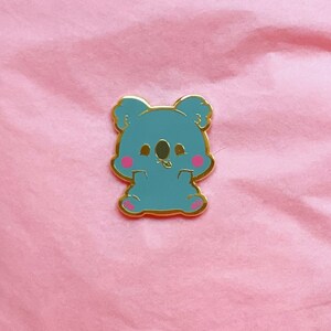 Puede incluir: Un pin de esmalte azul con un koala de dibujos animados con mejillas rosadas. El koala est&aacute; sentado con los brazos cruzados y tiene una peque&ntilde;a hoja en la boca.