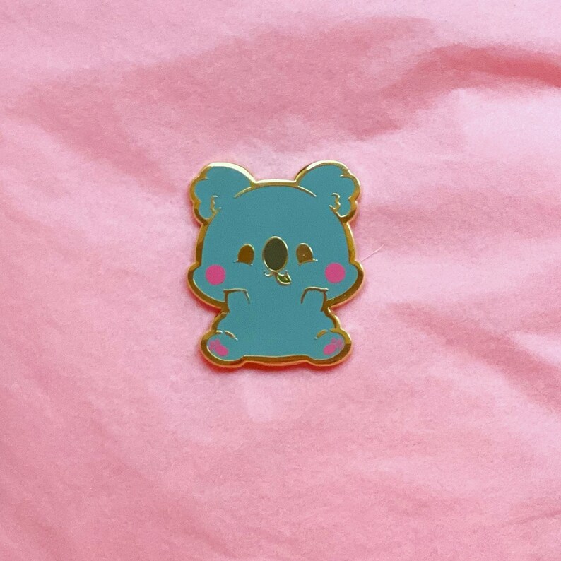 Puede incluir: Un pin de esmalte azul con un koala de dibujos animados con mejillas rosadas. El koala est&aacute; sentado con los brazos cruzados y tiene una peque&ntilde;a hoja en la boca.