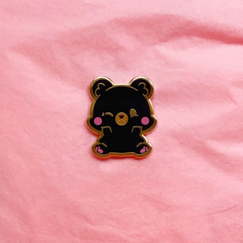 Puede incluir: Un pin de esmalte dorado con un oso negro con mejillas rosadas y un ojo gui&ntilde;ando. El oso est&aacute; sentado con las patas metidas delante de &eacute;l.