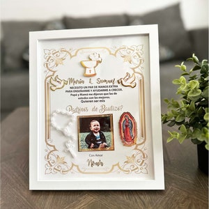 May include: A white frame with a gold border and a white background. The frame contains a photo of a baby boy, a rosary, a religious icon, and a text that reads "Maria & Samuel Necesito un par de manos extra para enseñarme y ayudarme a crecer. Papa y Mama me dijeron que las de ustedes son las mejores. Quieren ser mis Padrinos de Bautizo? Con Amor Noah".