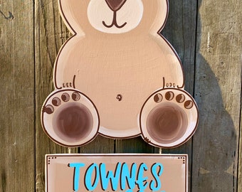 May include: A wooden bear cutout with a brown and tan color scheme. The bear is smiling and has a light brown belly. The bear is hanging from a string. Below the bear is a wooden sign with the name "Townes Cooper Lynn" written in blue paint.