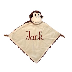 May include: A cream-coloured baby blanket with a brown border and a plush monkey head. The name "Jack" is embroidered in brown script on the blanket. The monkey has a tan face and brown fur.