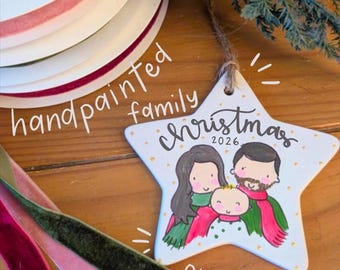 May include: A white star-shaped ceramic ornament featuring a hand-painted illustration of a family of three with the text "christmas 2026" in black script. The ornament includes a jute twine hanger and is accented with small gold painted dots.
