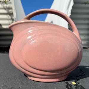 May include: A vintage, pink ceramic pitcher with a large handle. The pitcher has a rounded body with a spout and a curved handle. The exterior features a series of horizontal ridges. The pitcher is sitting on a dark surface.