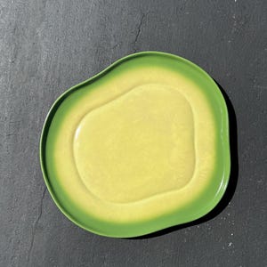 May include: A ceramic plate shaped like an avocado half. The plate features a light yellow center and a green rim, mimicking the fruit's colors. The plate is designed with a smooth, glossy finish.