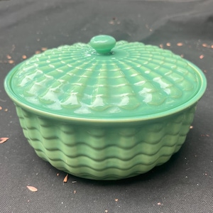 May include: A round, seafoam green ceramic serving dish with a lid. The lid has a textured, fan-like pattern, and a small, round knob. The dish's body features a wavy, basket-weave design. The dish is likely used for serving food.