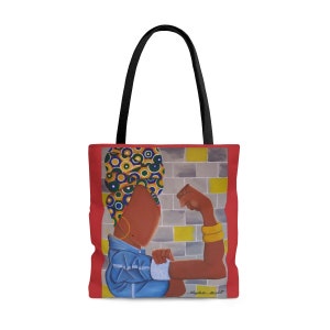 May include: Red tote bag with a colorful illustration of a woman with her fist raised in the air. The woman is wearing a blue shirt and a colorful head wrap. The background is a brick wall with yellow accents. The text "Nygela. M. Holt" is in the bottom right corner.