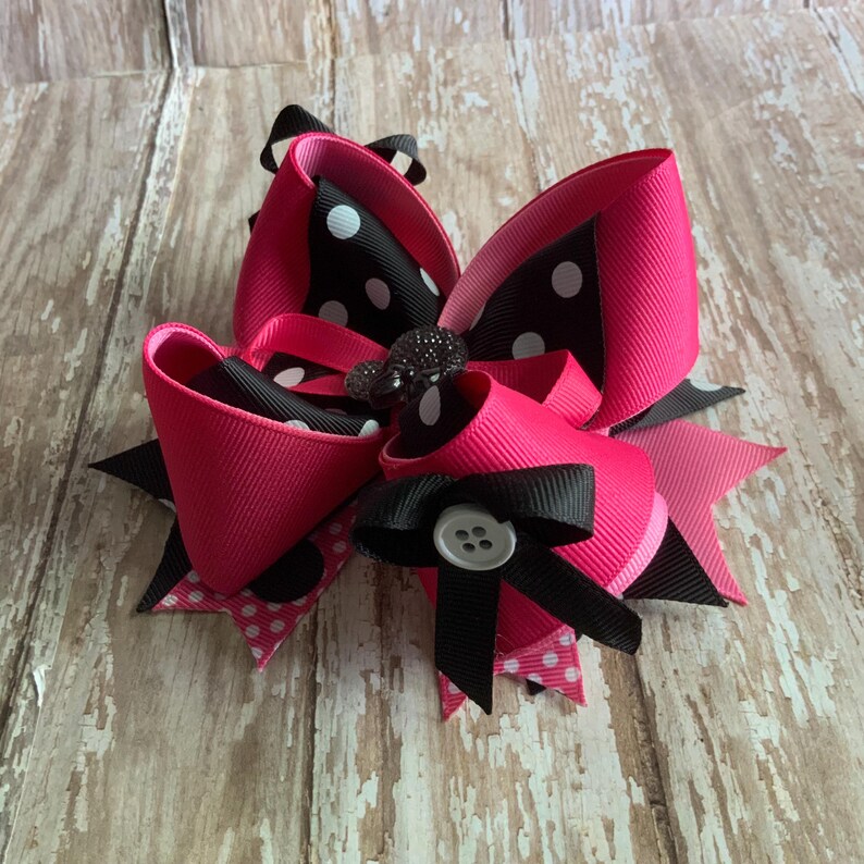 Op de afbeelding: Een grote, gelaagde haarstrik met helder roze en zwarte linten. De strik heeft een stippenpatroon en is versierd met een decoratieve knoop en een juwelen middenstuk. De strik staat tegen een houten achtergrond.
