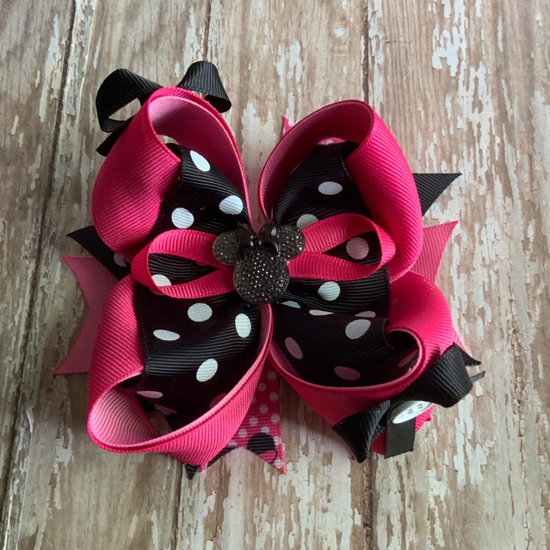 Op de afbeelding: Een grote haarstrik met helder roze en zwarte linten met witte stippen. Een zwart, glinsterend muisvormig ornament is gecentreerd op de strik. De strik staat tegen een houten achtergrond.