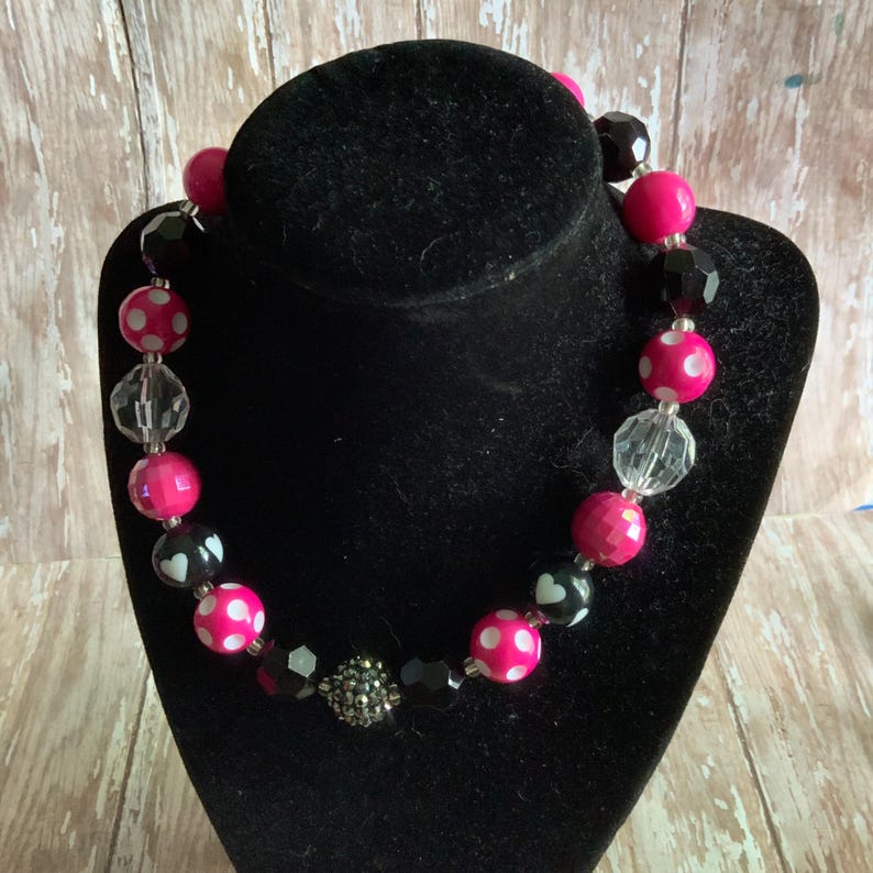 Op de afbeelding: Een opvallende ketting met roze, zwarte en transparante kralen. De roze kralen hebben witte stippen. De ketting wordt gepresenteerd op een zwarte buste. De kralen zijn rond en facetgeslepen, met een zilverkleurige accentkralen.