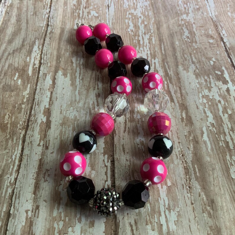 Op de afbeelding: Een ketting met roze, zwarte en transparante kralen. De kralen zijn rond, facetgeslepen en sommige hebben witte stippen of hartpatronen. De ketting ligt op een verweerd houten oppervlak.