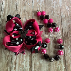 Op de afbeelding: Een roze en zwarte haarstrik met witte stippen en een strass Mickey Mouse-versiering. Een bijpassende kralenketting met roze, zwarte en transparante kralen is ook afgebeeld. De strik en de ketting liggen op een houten oppervlak.