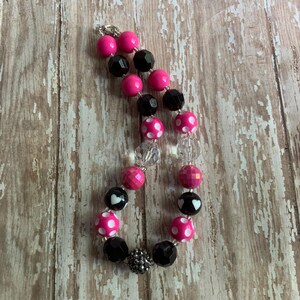 Op de afbeelding: Een ketting met roze, zwarte en transparante kralen. De ketting heeft effen roze kralen, facetgeslepen zwarte kralen en stippen kralen met witte stippen. Sommige kralen hebben een hartontwerp. De ketting ligt op een houten oppervlak.