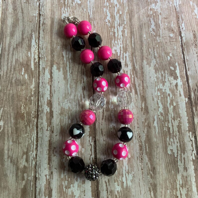 Op de afbeelding: Een ketting met roze, zwarte en transparante kralen. De ketting heeft effen roze kralen, facetgeslepen zwarte kralen en stippen kralen met witte stippen. Sommige kralen hebben een hartontwerp. De ketting ligt op een houten oppervlak.