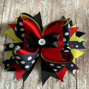 May include: A large, layered hair bow featuring red, black, yellow, and white polka dot ribbons. The bow has a button center and is set against a wooden background. The bow is a decorative hair accessory.