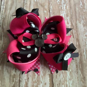 Op de afbeelding: Een grote, gelaagde haarstrik in felroze, zwart en wit gestippeld lint. De strik heeft een kleine zwarte strik bovenop, een decoratieve knoop en een strass Mickey Mouse-embleem in het midden. Ontworpen voor een kind.