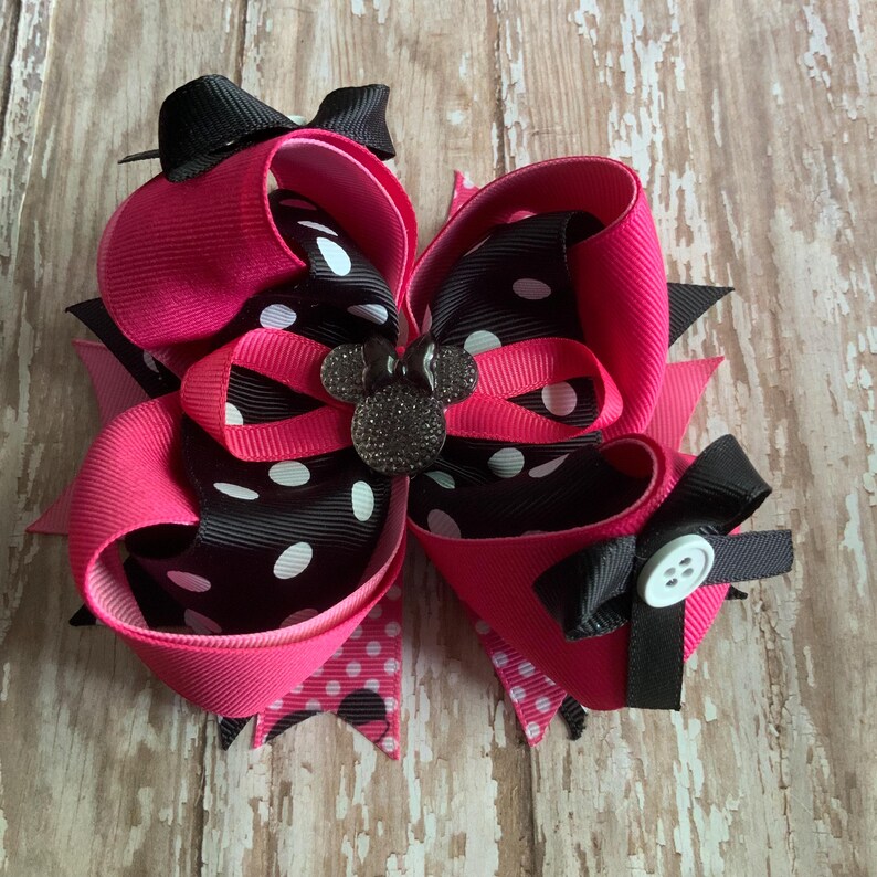 Op de afbeelding: Een grote, gelaagde haarstrik in felroze, zwart en wit gestippeld lint. De strik heeft een kleine zwarte strik bovenop, een decoratieve knoop en een strass Mickey Mouse-embleem in het midden. Ontworpen voor een kind.