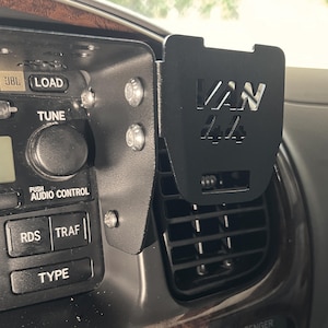 Peut inclure: Un support de téléphone de voiture noir avec le texte "VAN" imprimé dessus. Le support est fixé à l'aération d'une voiture.