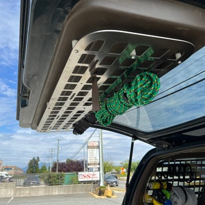 May include: A silver metal storage rack mounted inside a vehicle's rear hatch. A green and yellow rope is coiled and secured to the rack. The vehicle's rear window is visible, with a view of a street and buildings.