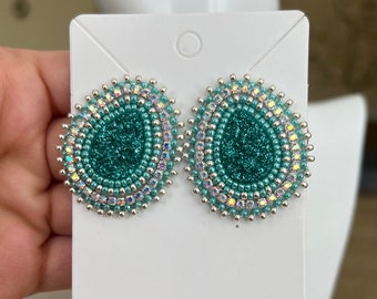 May include: Teal and silver beaded earrings with a glitter center. The earrings are round and have a beaded design.