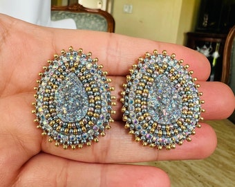 May include: A pair of silver and gold oval-shaped earrings with a teardrop-shaped iridescent stone in the center. The earrings are beaded with silver and gold beads.
