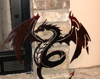 Puede incluir: Una pieza de arte mural de metal oscuro con forma de dragón, con alas grandes y extendidas, una cola larga y curva que termina en punta, y detalles recortados en el cuerpo, con un acabado en degradado de color rojo oscuro y negro.